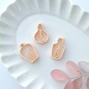 May include: Three plastic cookie cutters shaped like bottles with a sprig of greenery inside. The cutters are on a white plate with a scalloped edge.