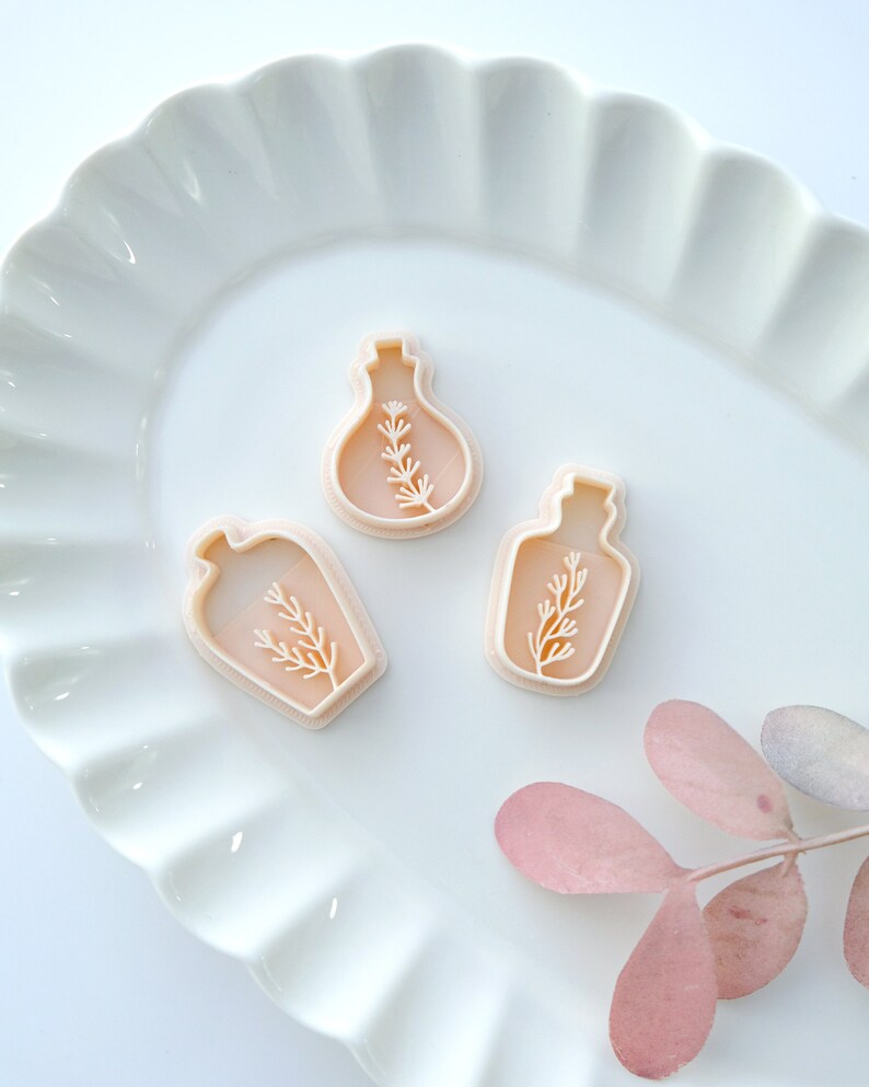 May include: Three plastic cookie cutters shaped like bottles with a sprig of greenery inside. The cutters are on a white plate with a scalloped edge.