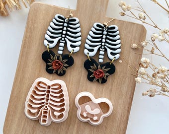 May include: A pair of skeleton ribcage earrings with a black and white design and a red rose detail. The earrings are shown with a matching skeleton ribcage and rose cookie cutter.