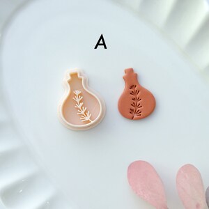 May include: A white plastic mold and a terracotta clay impression of a small bottle with a sprig of leaves inside. The mold and impression are on a white plate with the letter "A" printed on the plate.