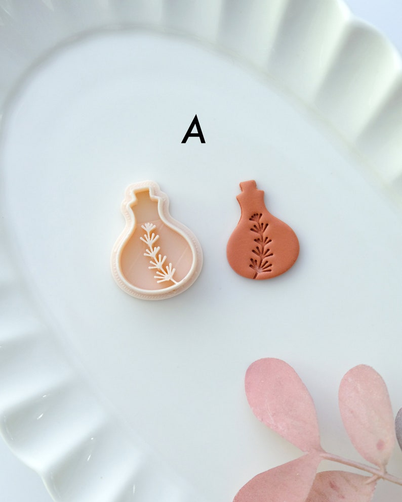 May include: A white plastic mold and a terracotta clay impression of a small bottle with a sprig of leaves inside. The mold and impression are on a white plate with the letter "A" printed on the plate.