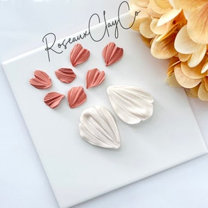 May include: A set of polymer clay flower petals in two colors: white and a light peachy pink. The petals are arranged on a white surface. The text "Rosseaux Clay Co." is visible in the top center of the image.