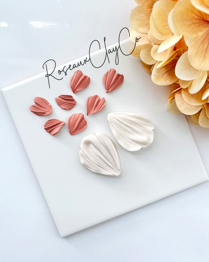 May include: A set of polymer clay flower petals in two colors: white and a light peachy pink. The petals are arranged on a white surface. The text "Rosseaux Clay Co." is visible in the top center of the image.