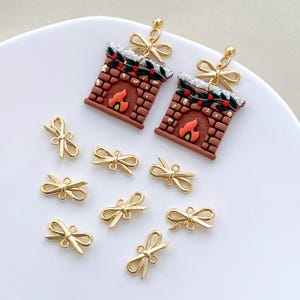 以下が含まれることがあります： ミニチュアレンガの暖炉と金色のリボンが特徴のクリスマスをテーマにしたイヤリング。追加の金色のリボンチャームも付いています。イヤリングは、お祝いのホリデーデザインです。