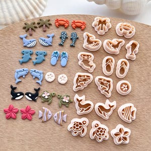 Könnte beinhalten: Ein Set mit 20 Ausstechern in verschiedenen Designs zum Thema Meer, darunter Palmen, Wale, Haie, Schildkröten, Fische, Muscheln und Flip-Flops. Die Ausstecher sind aus hellbeigefarbenem Kunststoff.