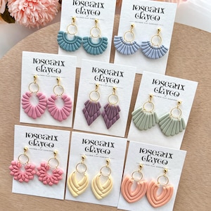 May include: A collection of polymer clay earrings in various shapes and colors, including blue, purple, pink, yellow, and green. Each pair features gold-colored hoops and is displayed on a white card with the text "loseaux clayco".