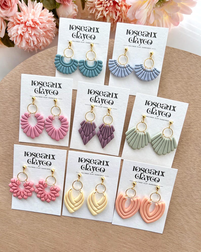May include: A collection of polymer clay earrings in various shapes and colors, including blue, purple, pink, yellow, and green. Each pair features gold-colored hoops and is displayed on a white card with the text "loseaux clayco".