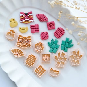 Puede incluir: Un conjunto de 17 cortadores de galletas de plástico en varias formas y colores, incluyendo una danza de leones, un abanico, una linterna, una bolsa de dinero y bambú. Los cortadores están dispuestos en un plato blanco.