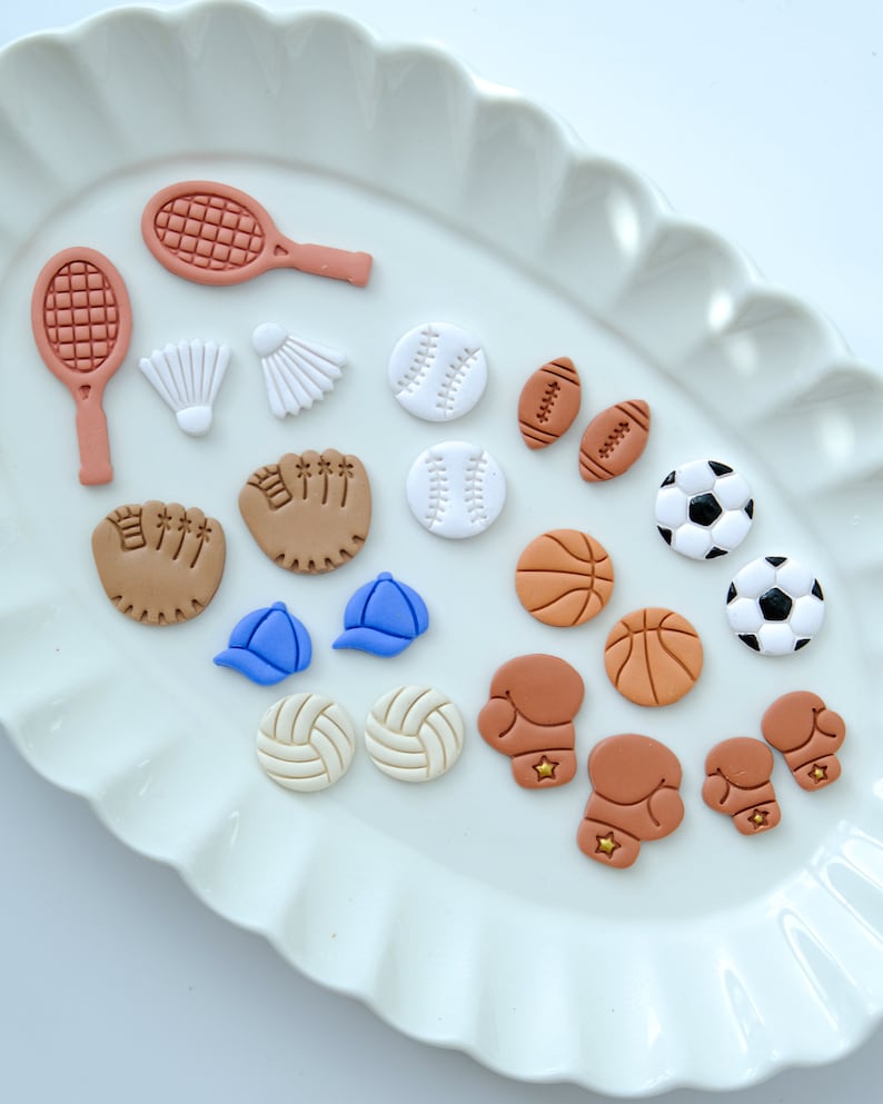 Pu&ograve; includere: Un piatto bianco con una variet&agrave; di ciondoli in argilla a tema sportivo, tra cui palle da baseball, palloni da basket, palloni da calcio, palloni da calcio, palloni da pallavolo, racchette da tennis, racchette da badminton, guantoni da boxe e guanti da baseball.