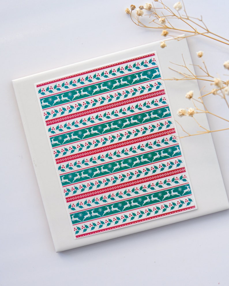 May include: White background with a sheet of wrapping paper featuring a repeating pattern of red berries, green holly leaves, white snowflakes, and teal reindeer.