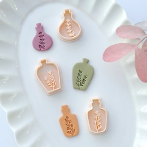 May include: Five different cookie cutters in the shape of bottles with a sprig of leaves inside. The cutters are made of white plastic and are arranged on a white plate with a scalloped edge.