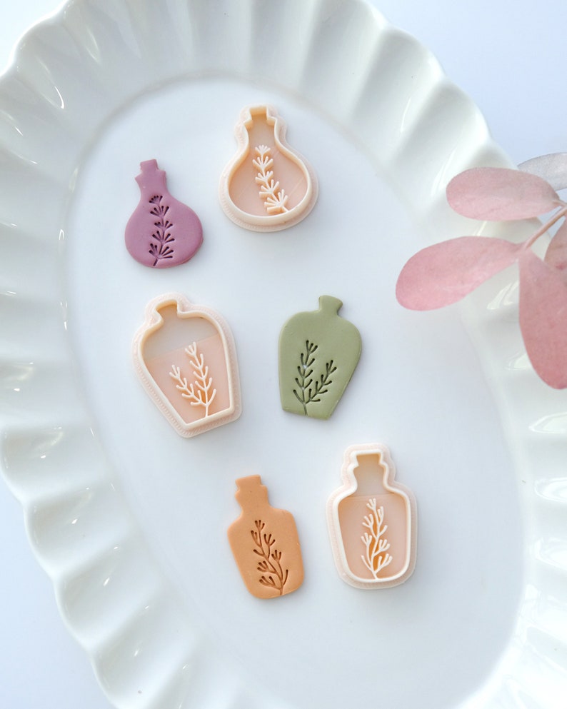 May include: Five different cookie cutters in the shape of bottles with a sprig of leaves inside. The cutters are made of white plastic and are arranged on a white plate with a scalloped edge.
