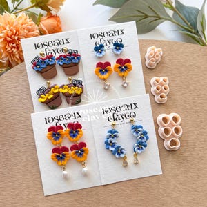 May include: Handmade polymer clay earrings in pansy flower designs. The earrings feature blue, yellow, and red colours, with some designs including pearl accents and rhinestone details. The brand name "Roseaux Clayco" is visible on the cards. The earrings are presented on white cards.