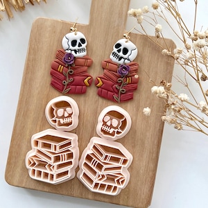 May include: Skull-shaped earrings with book stacks, purple flowers, and gold hooks. Also, two skull and two book-shaped cookie cutters. The items are displayed on a wooden chopping board. The earrings and cutters are in shades of red and brown.
