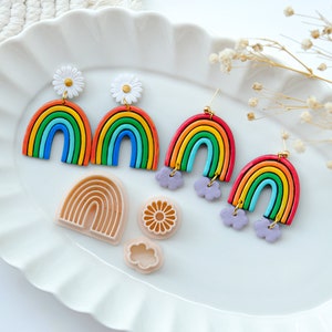 May include: Three pairs of rainbow earrings with white daisy charms and purple cloud charms.  The earrings are displayed on a white plate with rainbow and cloud shaped cookie cutters.