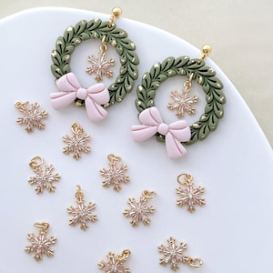 Op de afbeelding: Oorbellen met groene kransontwerpen, gouden details en roze strikken. Elke oorbel heeft een bungelende sneeuwvlokbedel. Extra sneeuwvlokbedels zijn verspreid rond de oorbellen.