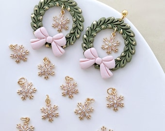 May include: Earrings featuring green wreath designs with gold accents and pink bows. Each earring has a dangling snowflake charm. Additional snowflake charms are scattered around the earrings, all with gold-colored findings.
