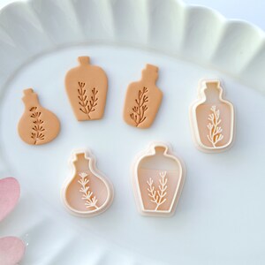 May include: Five cookie cutters in the shape of bottles with a sprig of leaves inside. The cutters are on a white plate with a scalloped edge.