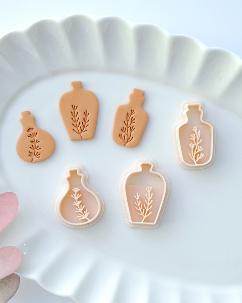 May include: Five cookie cutters in the shape of bottles with a sprig of leaves inside. The cutters are on a white plate with a scalloped edge.