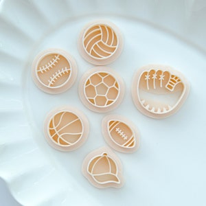 Pu&ograve; includere: Sette tagliabiscotti di plastica a forma di attrezzatura sportiva, tra cui una palla da baseball, una palla da basket, un pallone da football americano, un pallone da calcio, una palla da pallavolo, un berretto da baseball e un guanto da baseball. I tagliabiscotti sono su un piatto bianco.