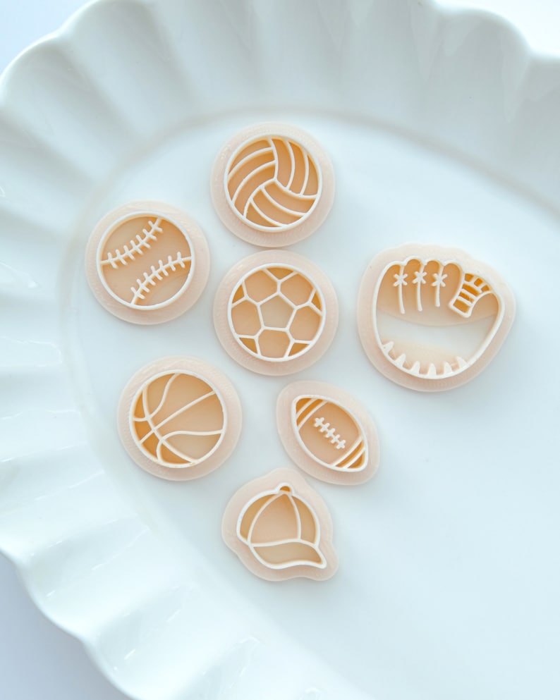 Pu&ograve; includere: Sette tagliabiscotti di plastica a forma di attrezzatura sportiva, tra cui una palla da baseball, una palla da basket, un pallone da football americano, un pallone da calcio, una palla da pallavolo, un berretto da baseball e un guanto da baseball. I tagliabiscotti sono su un piatto bianco.