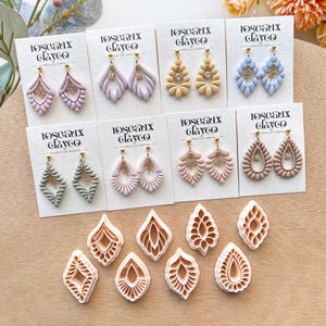 Puede incluir: Una colección de pendientes de arcilla polimérica en varios colores, incluyendo rosa, beige y azul, exhibidos en tarjetas blancas. Los pendientes presentan diseños intrincados y se combinan con cortadores de arcilla a juego.