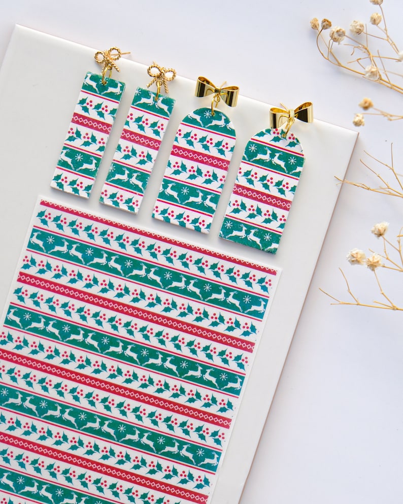 May include: Four Christmas-themed earrings with a white background and green and red patterns. The earrings are shaped like rectangular tags with a gold bow at the top. The pattern features a repeating design of holly leaves, red berries, and reindeer.