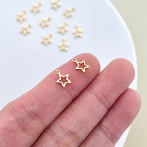 Puede incluir: Primer plano de pequeños dijes de estrella dorados. Los dijes tienen forma de estrella hueca con un pequeño lazo en la parte superior para sujetarlos a las joyas. Varios otros dijes de estrella están dispersos en el fondo.