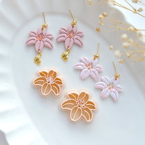 May include: Four pairs of handmade polymer clay earrings featuring a lily flower design. Two pairs are pink with gold accents, one pair is white with gold accents, and one pair is a light peach color.