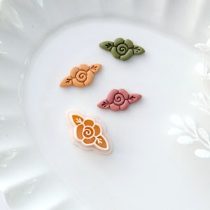 May include: Four small, decorative floral embellishments in various shades of orange, green, and pink. Each embellishment features a stylized rose with leaves.