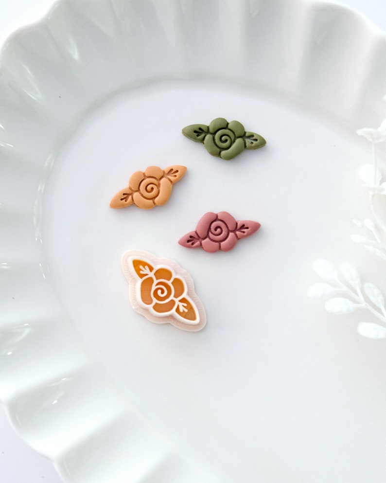 May include: Four small, decorative floral embellishments in various shades of orange, green, and pink. Each embellishment features a stylized rose with leaves.
