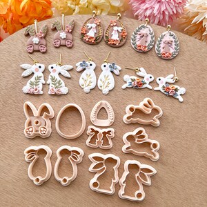 May include: A set of 14 polymer clay bunny and egg earrings and cookie cutters. The earrings feature white bunnies with floral accents and brown eggs with floral designs. The cookie cutters are in the shape of bunnies and eggs.