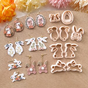 May include: A set of 10 polymer clay bunny and egg-shaped earrings and cookie cutters. The earrings are handmade and feature pastel pink, white, and gold accents. The cookie cutters are made of metal and are designed to cut out bunny and egg shapes.