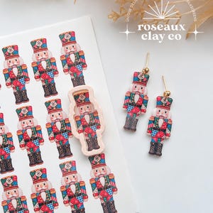 May include: A sheet of nutcracker-themed silkscreen transfers featuring multiple colorful nutcracker illustrations, a single beige nutcracker-shaped clay cutter, and a pair of dangling nutcracker earrings with gold-toned posts.