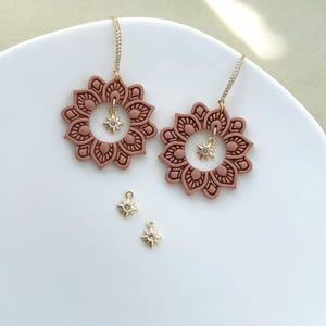 May include: Two pairs of earrings and two star-shaped charms. The earrings feature a floral design in a muted rose color, with a gold-colored chain and a star-shaped charm. The charms are gold-colored with a clear crystal in the center.