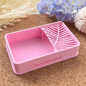 Puede incluir: Un organizador rectangular rosa claro con un escurridor con diseño de hojas. El organizador tiene un borde elevado y el texto "roseandclayco" está impreso en el lateral. El organizador está sobre una superficie de corcho con flores en el fondo.