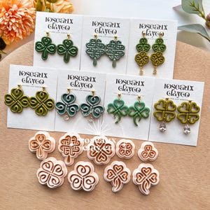 Può includere: Una collezione di orecchini a forma di trifoglio verde e taglierini in argilla abbinati. Gli orecchini sono esposti su cartoncini bianchi con perni dorati. Gli orecchini sono disponibili in varie tonalità di verde e presentano diversi design. I taglierini in argilla sono marroni chiari.
