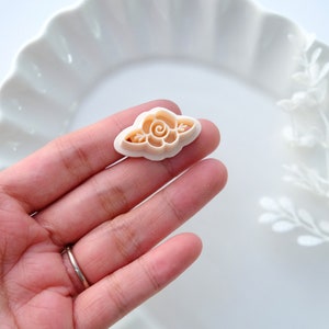 May include: A white plastic cookie cutter shaped like a rose with a swirl in the center. The cutter is on a hand.