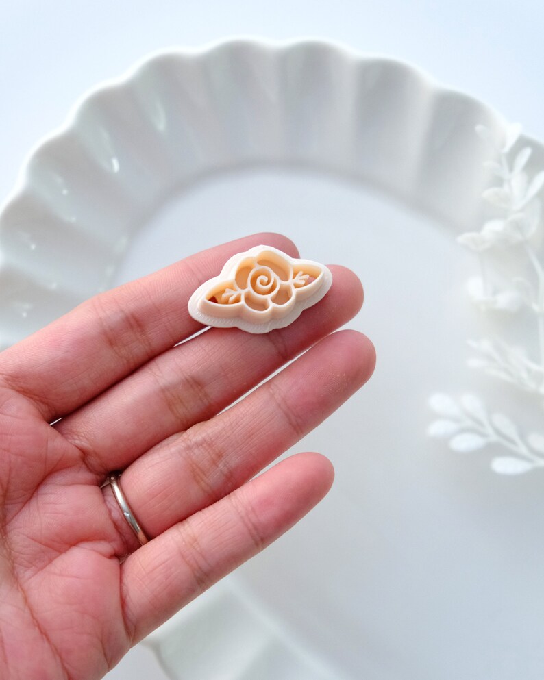 May include: A white plastic cookie cutter shaped like a rose with a swirl in the center. The cutter is on a hand.