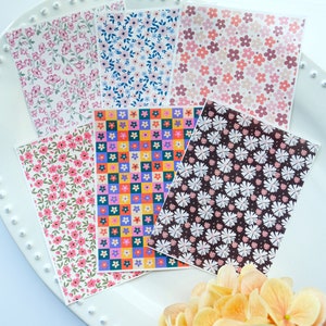 May include: Six sheets of patterned paper featuring floral designs. The patterns include pink and white flowers on a white background, blue and white flowers on a white background, pink and white flowers on a light pink background, colourful flowers in a checkered pattern, white flowers on a dark brown background, and pink and white flowers on a white background.