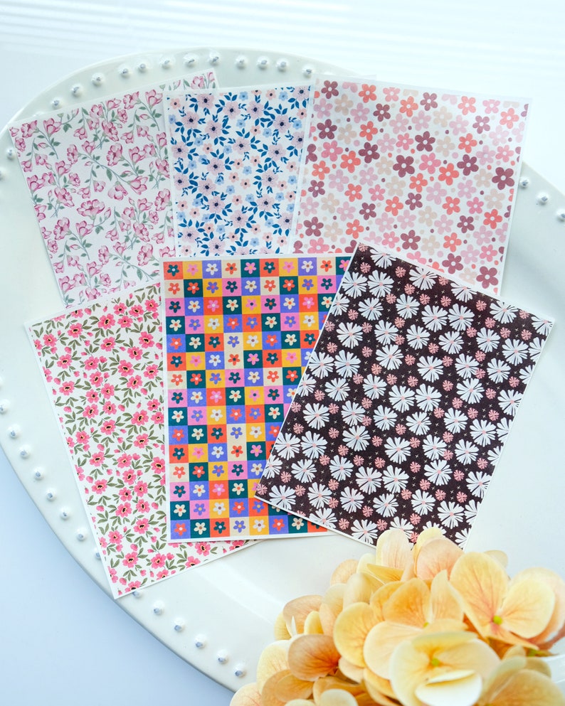 May include: Six sheets of patterned paper featuring floral designs. The patterns include pink and white flowers on a white background, blue and white flowers on a white background, pink and white flowers on a light pink background, colourful flowers in a checkered pattern, white flowers on a dark brown background, and pink and white flowers on a white background.