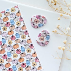May include: Two white ceramic pumpkin-shaped earrings with a colorful pumpkin pattern. The earrings are on a white surface with dried flowers.