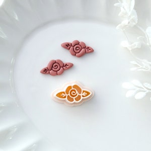 May include: Three polymer clay rose embellishments. Two are a deep pink color and one is a light pink with a yellow center.