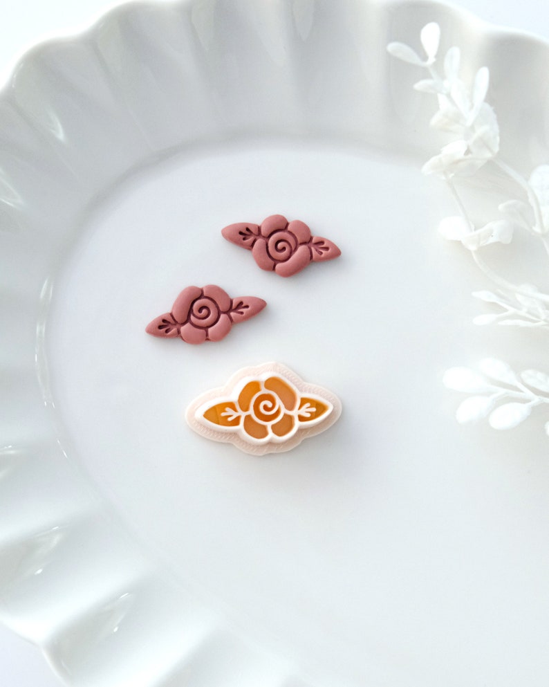 May include: Three polymer clay rose embellishments. Two are a deep pink color and one is a light pink with a yellow center.