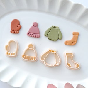 May include: Set of eight cookie cutters in the shape of winter clothing items. The cutters are made of white plastic and include a mitten, a hat, a sweater, a sock, and three additional cutters in the same shapes.
