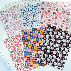 May include: Six sheets of patterned paper featuring floral designs. The patterns include pink and white flowers, blue and white flowers, pink and red flowers, colourful squares with flowers, white flowers on a black background, and pink and white flowers on a white background.