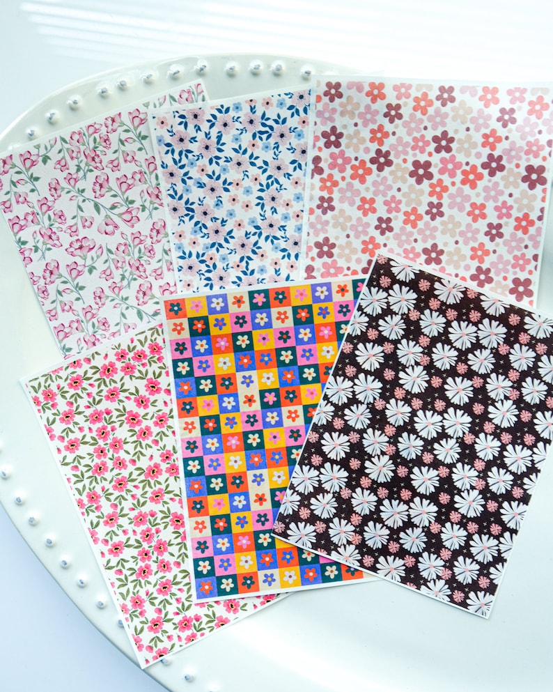 May include: Six sheets of patterned paper featuring floral designs. The patterns include pink and white flowers, blue and white flowers, pink and red flowers, colourful squares with flowers, white flowers on a black background, and pink and white flowers on a white background.