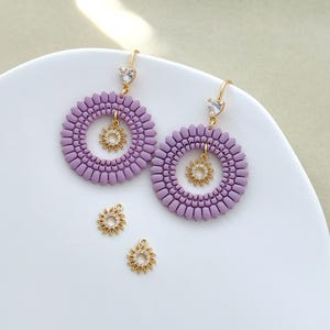 May include: A pair of lavender earrings with a circular design and a small gold sunburst charm in the center. The earrings have gold-colored hooks with a small, clear heart-shaped gem. Two additional sunburst charms are also present.