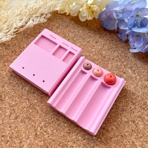 May include: Two pink silicone molds for making resin beads. The larger mold has three square sections labeled 10mm, 11mm, and 12mm. The smaller mold has four long, narrow sections. There are five pink resin beads in the smaller mold.