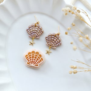 May include: A pair of delicate earrings featuring a seashell design with a gold star charm. The earrings are made of polymer clay and have a light pink and purple colour.